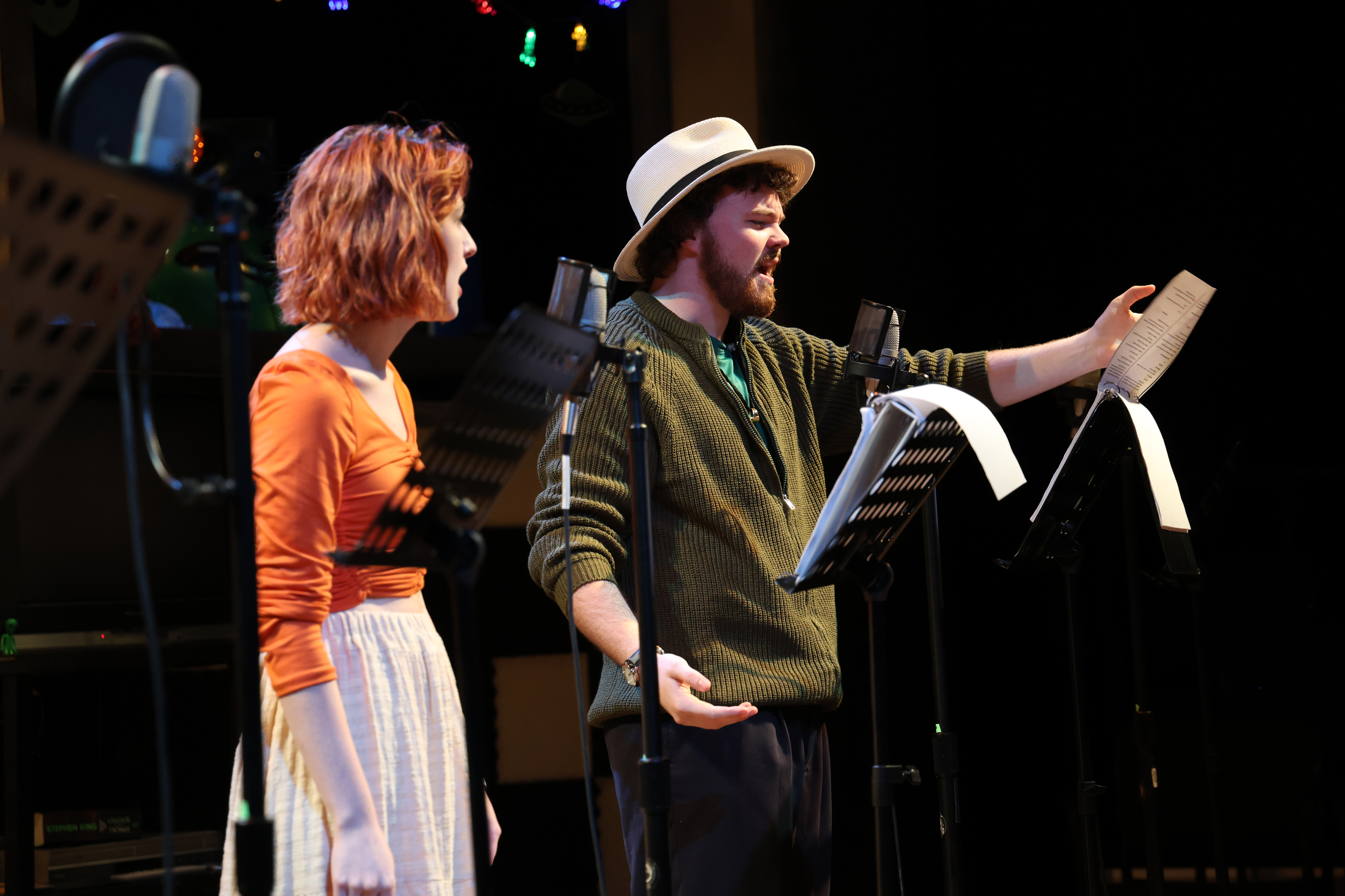 Two students on stage with radio scripts and mics in front of them from the production, Welcome to Our Village Please Invade Carefully