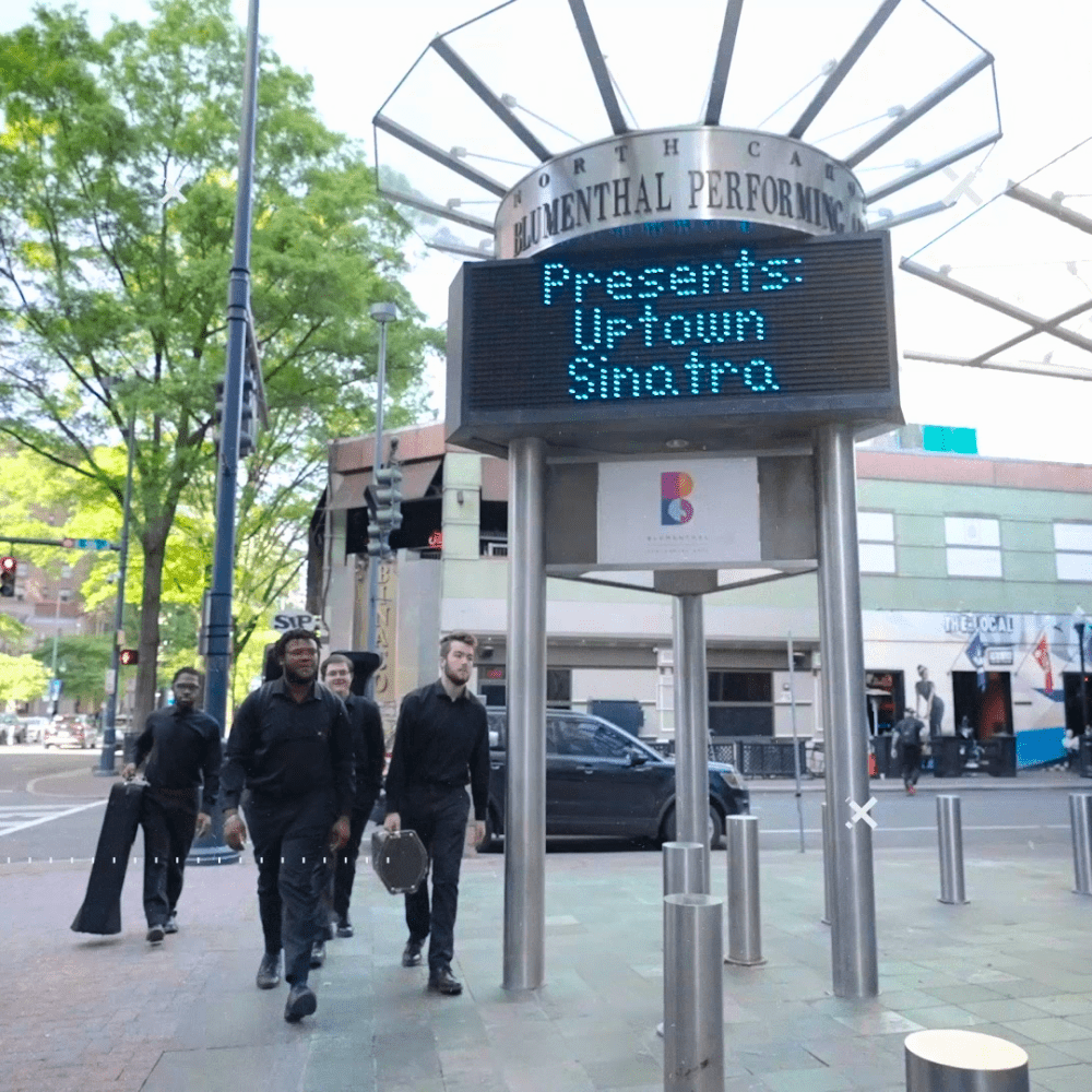 Music students walking through uptown with their instruments passing a sign that says performance tonight